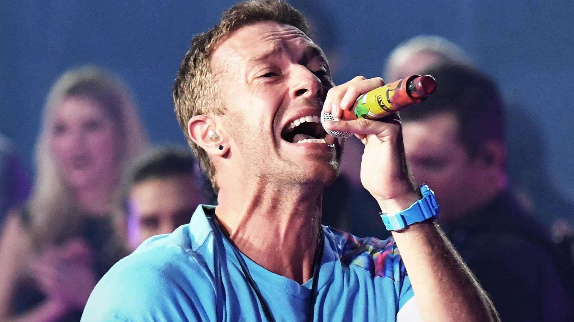 Chris Martin mengenakan kaos biru sedang menyanyikan lagu dalam sebuah konser